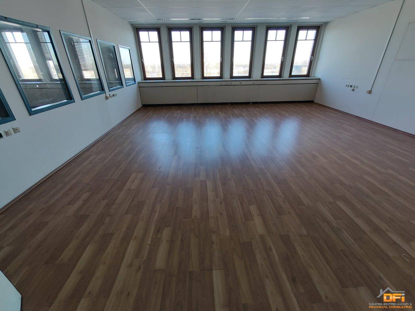 Großes Büro mit hellem Laminatboden und einer Fensterfront, die den Raum gut belichtet und eine Aussicht bietet.