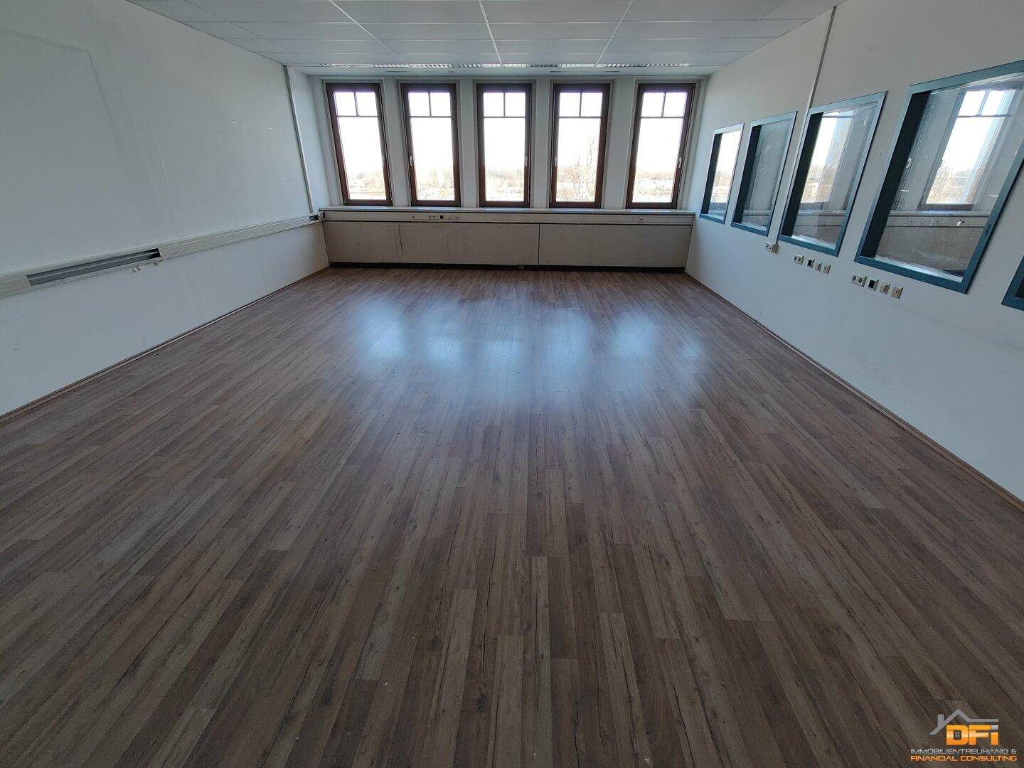 Großes Büro mit hellem Laminatboden und einer Fensterfront, die den Raum gut belichtet und eine Aussicht bietet.