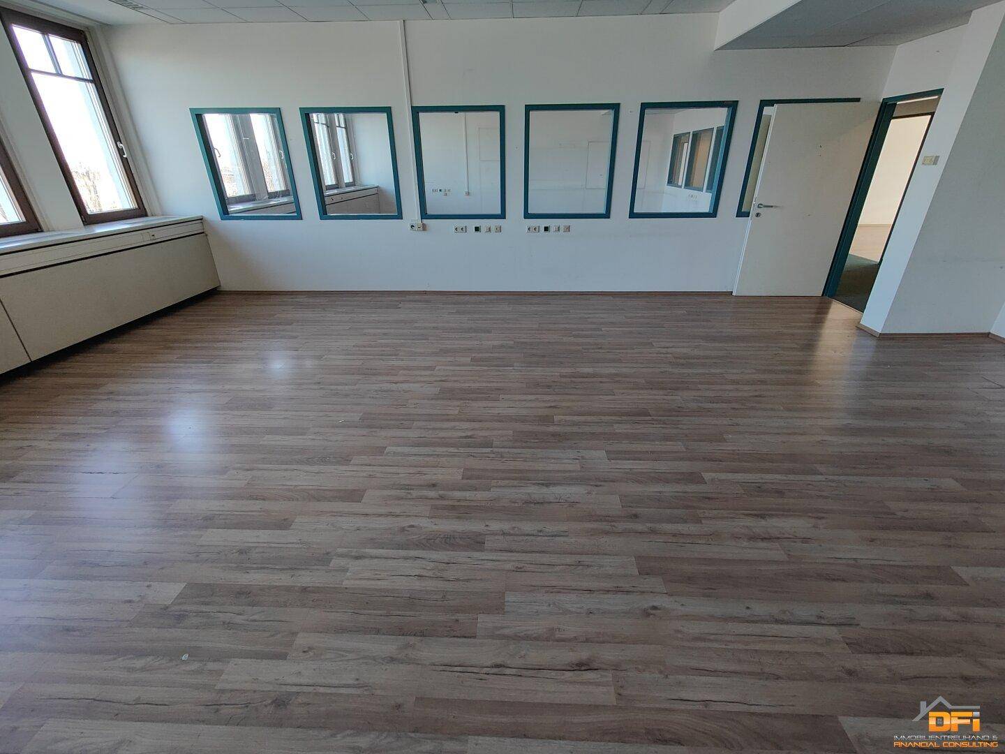 Geräumiges Büro mit hellem Laminatboden und mehreren Fenstern, die den Raum erhellen und eine Aussicht bieten.