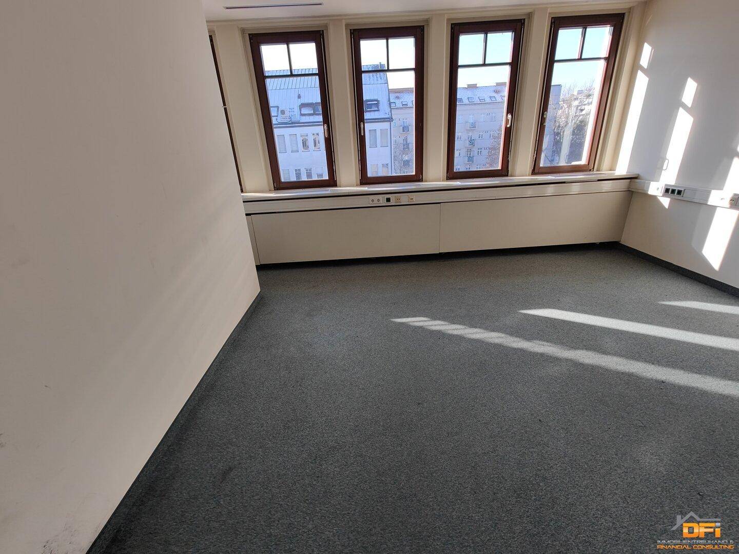 Heller Büroraum mit grauem Teppichboden und mehreren Fenstern, die einen Stadtblick bieten.