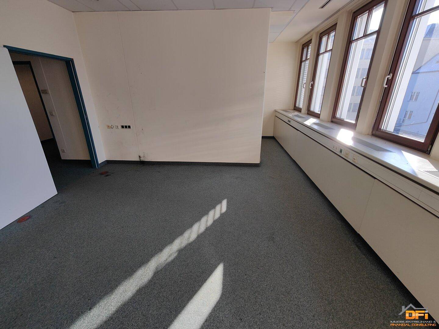 Geräumiger Büroraum mit grauem Teppichboden, großen Fenstern und viel Tageslichteinfall.
