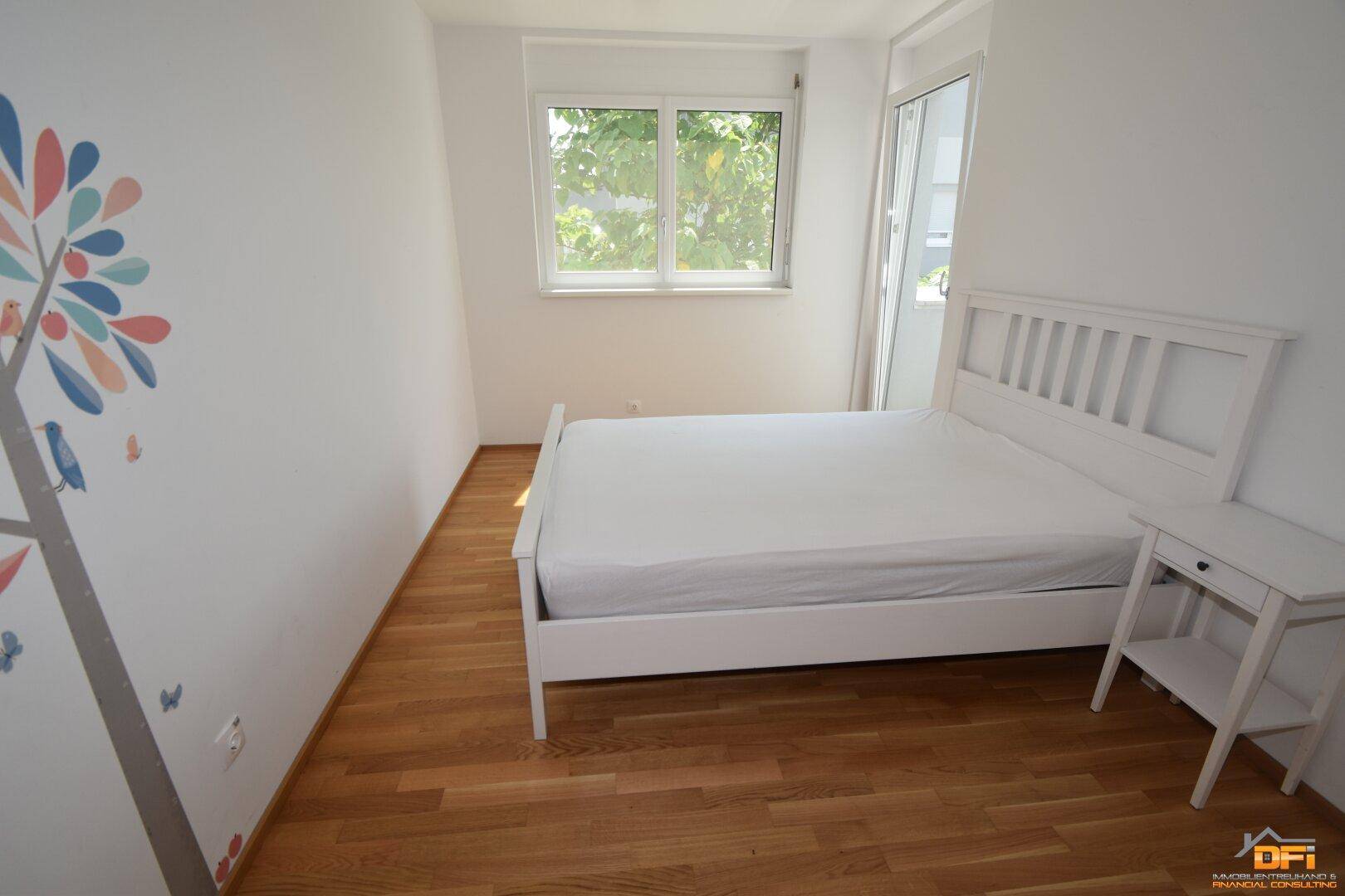 Helles Schlafzimmer mit Holzboden, weißem Bett und Fenster mit Blick ins Grüne.