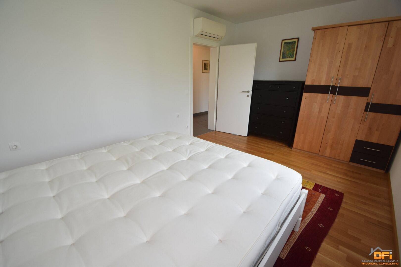 Schlafzimmer mit Holzboden, weißem Bett und großem Holzschrank, ausgestattet mit Klimaanlage.