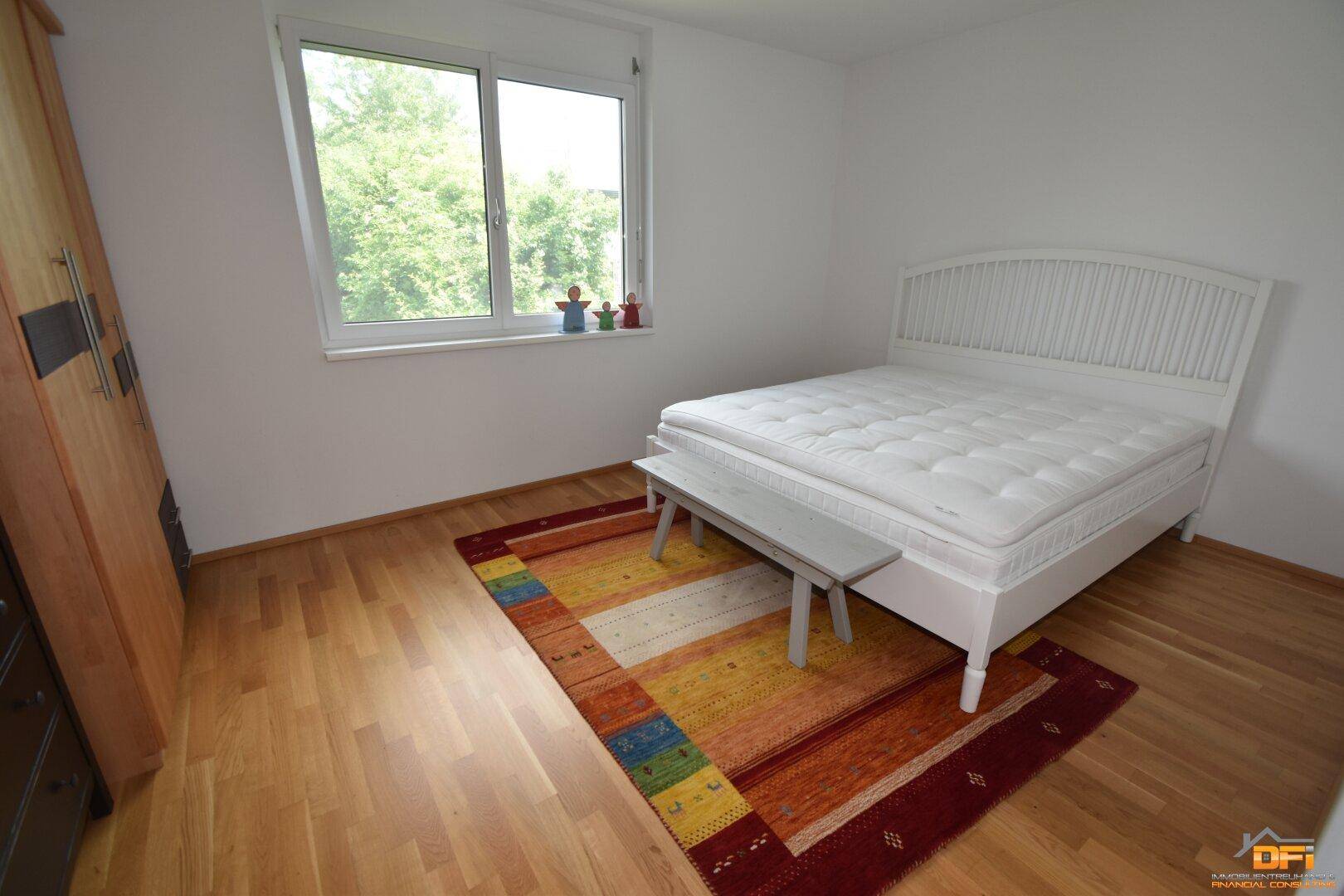 Schlafzimmer mit Holzboden, weißem Bett und farbenfrohem Teppich, bietet einen Blick ins Grüne.