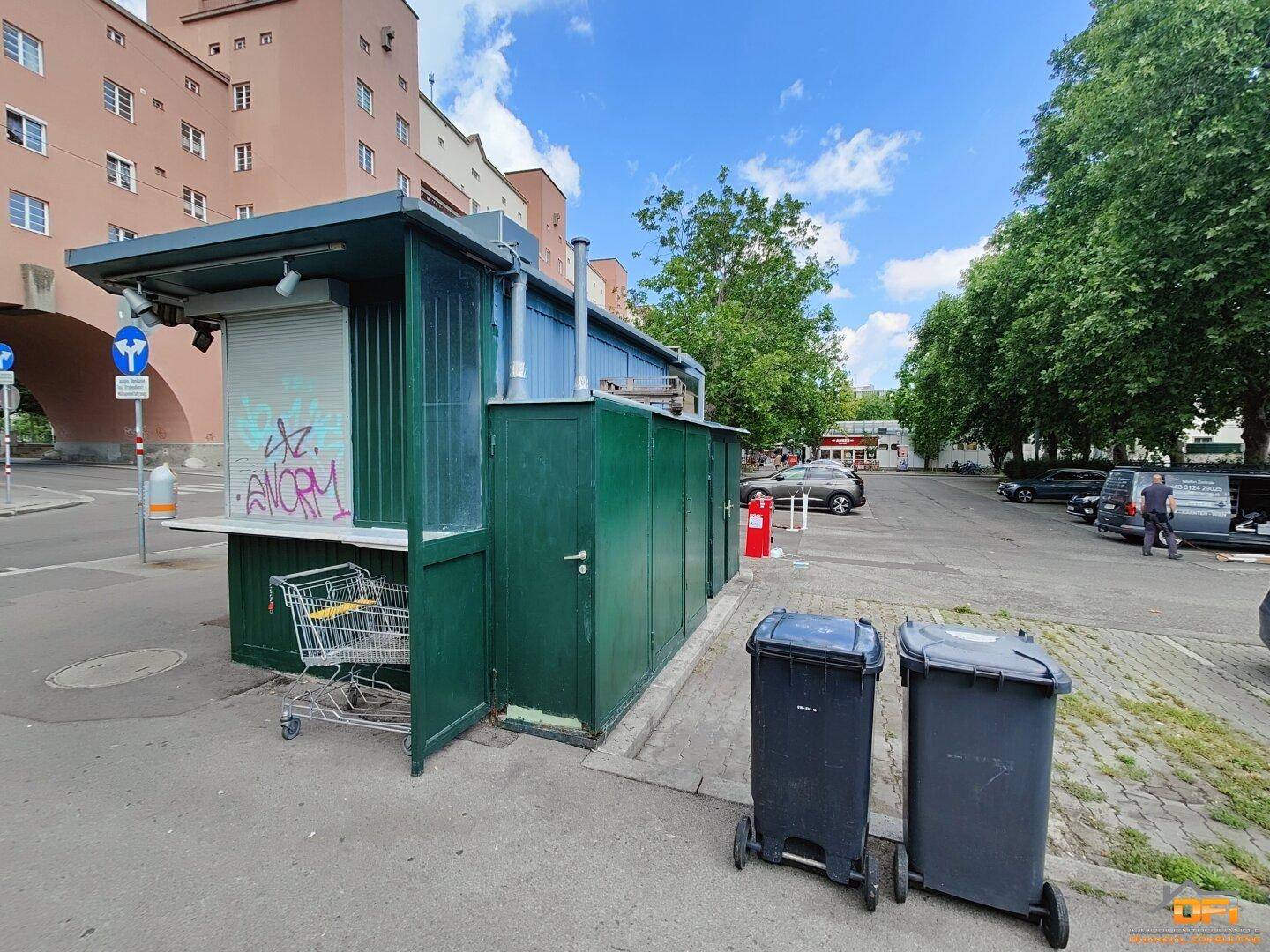 Rückseite des Imbissstandes mit Tür und Mülltonnen, im Hintergrund ein großes Wohngebäude und Parkplätze.