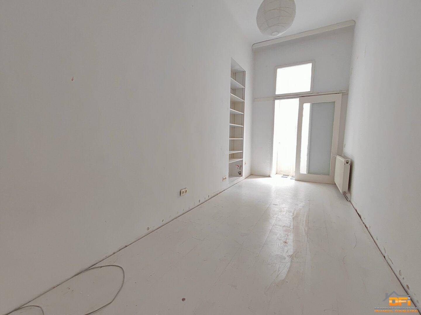 Schmaler, heller Raum mit weißem Holzboden und Zugang zu einem Balkon.