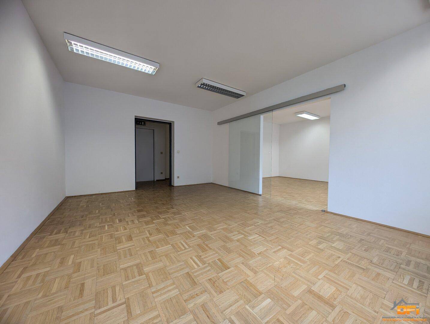 Helles Büro mit Parkettboden und Glasschiebetür, die zu einem weiteren Raum führt.
