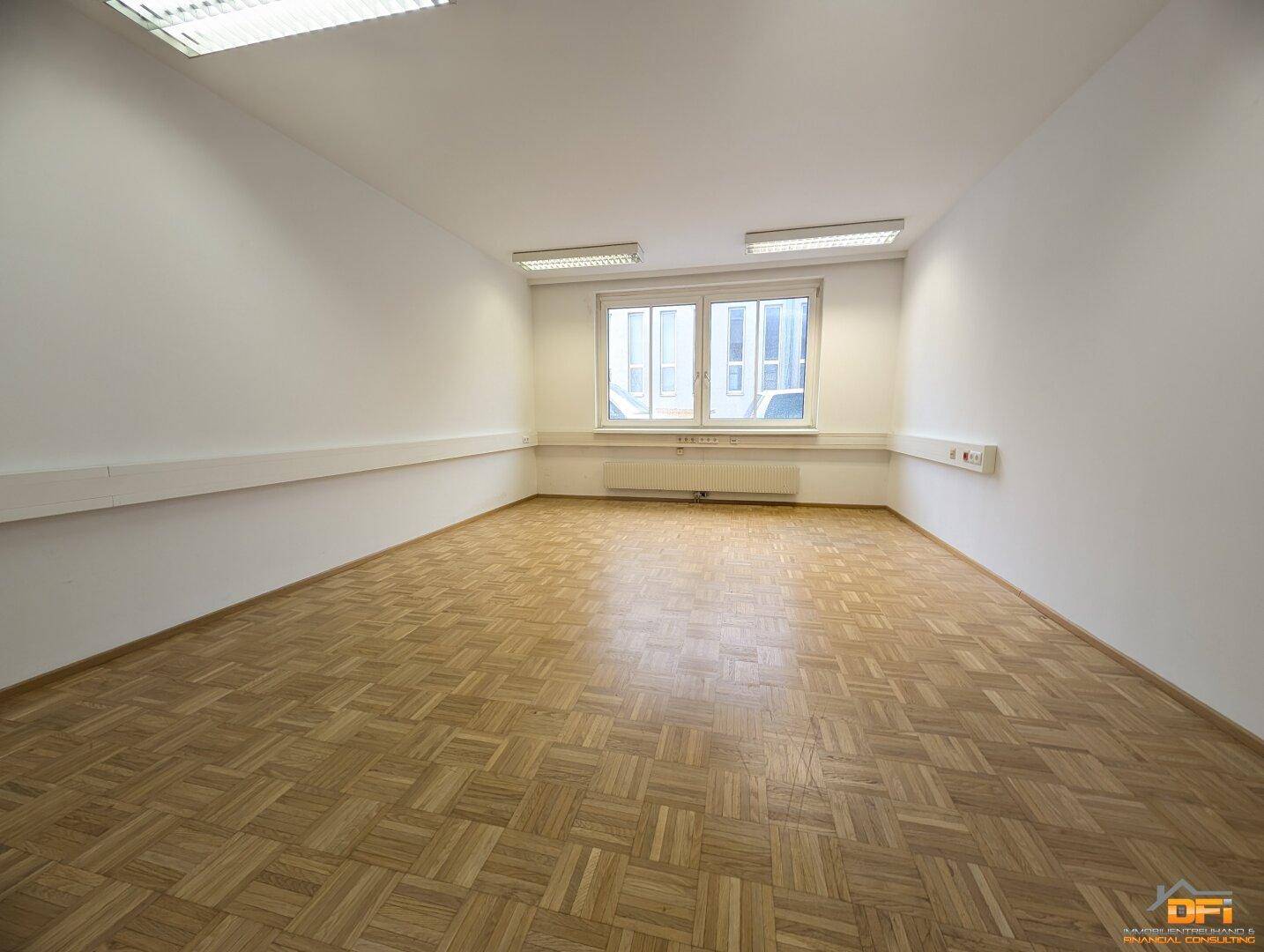 Leeres Büro mit Parkettboden und zwei großen Fenstern, die viel Tageslicht hereinlassen.