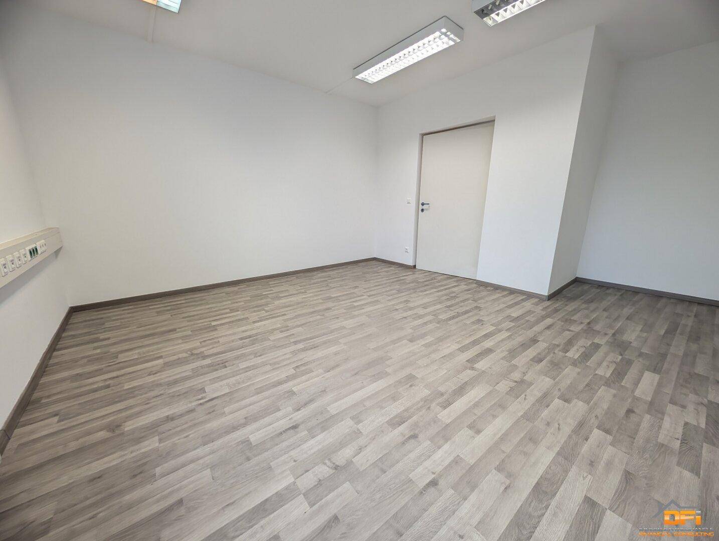 Geräumiger Büroraum mit hellem Holzoptikboden, weißen Wänden und effizienter Deckenbeleuchtung.