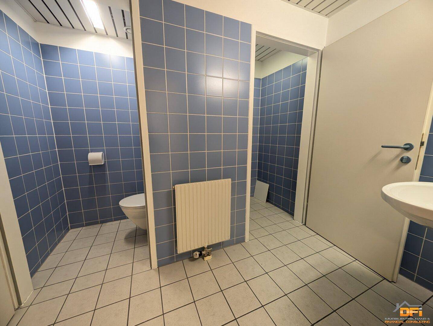 Öffentliche Toilette mit blauen Wandfliesen, weißem Fliesenboden, WC-Kabinen und Waschbecken.