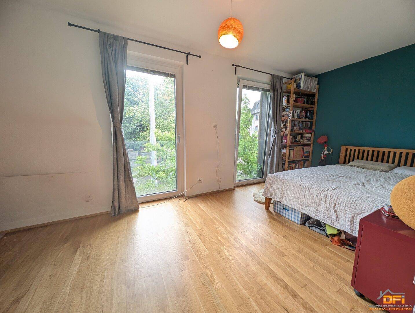 Schlafzimmer mit Holzboden, tealfarbener Akzentwand und großen Fenstern mit Blick ins Grüne.