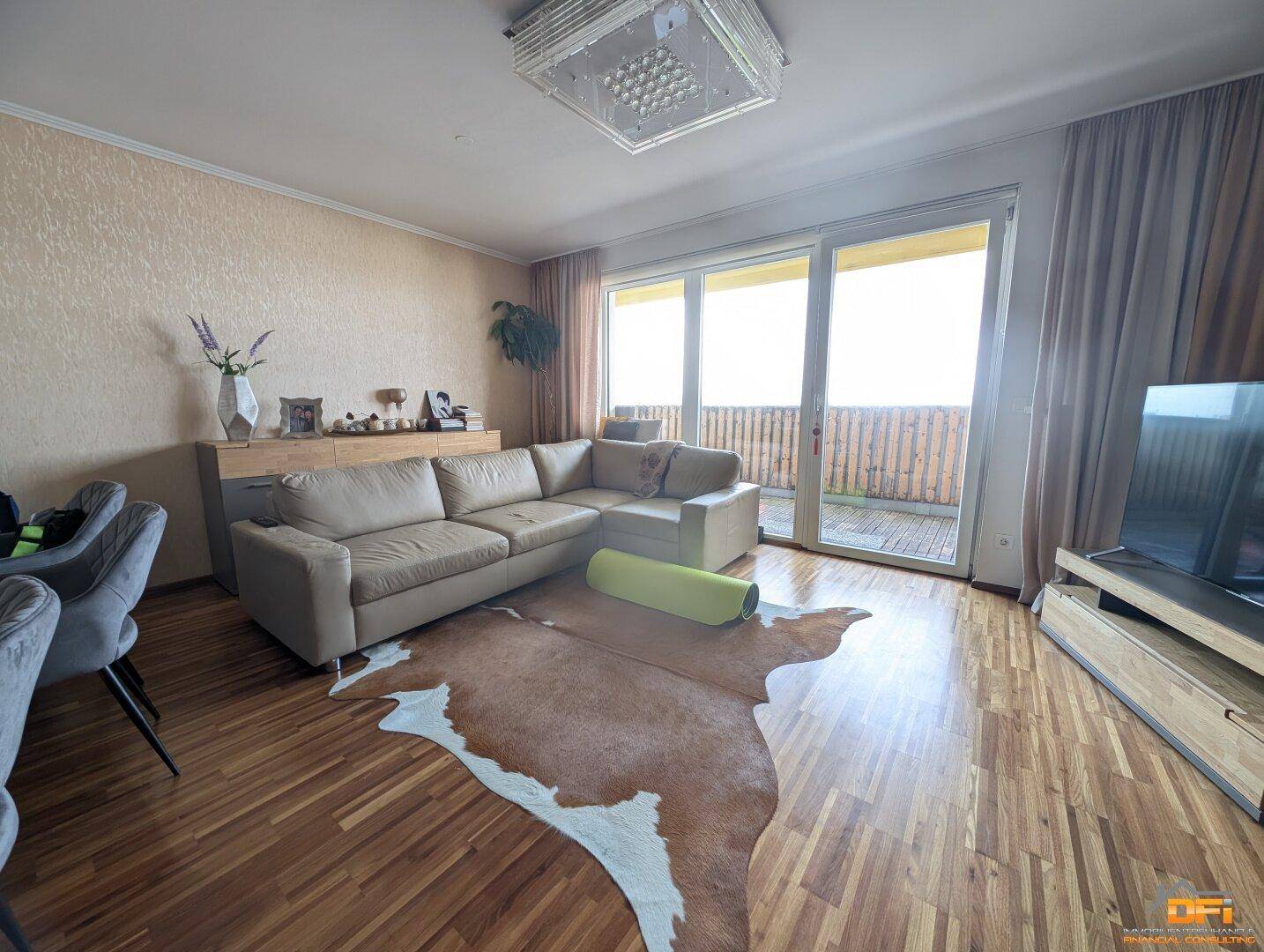 Gemütliches Wohnzimmer mit beigem Ecksofa, Holzboden und großen Fenstern zum Balkon.