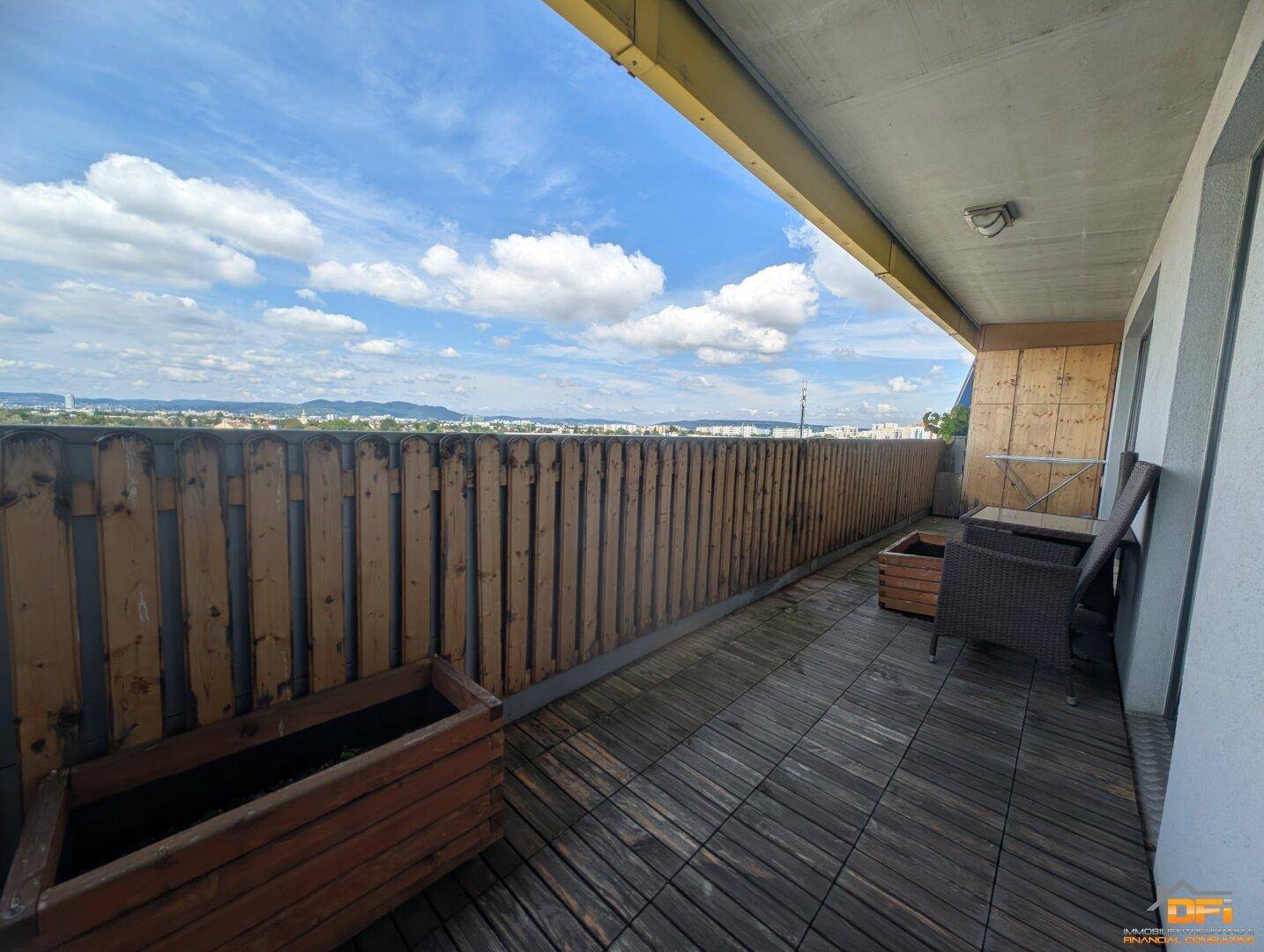 Langer Balkon mit Holzboden, Sitzgelegenheiten und weitem Blick über die Stadt und Berge.