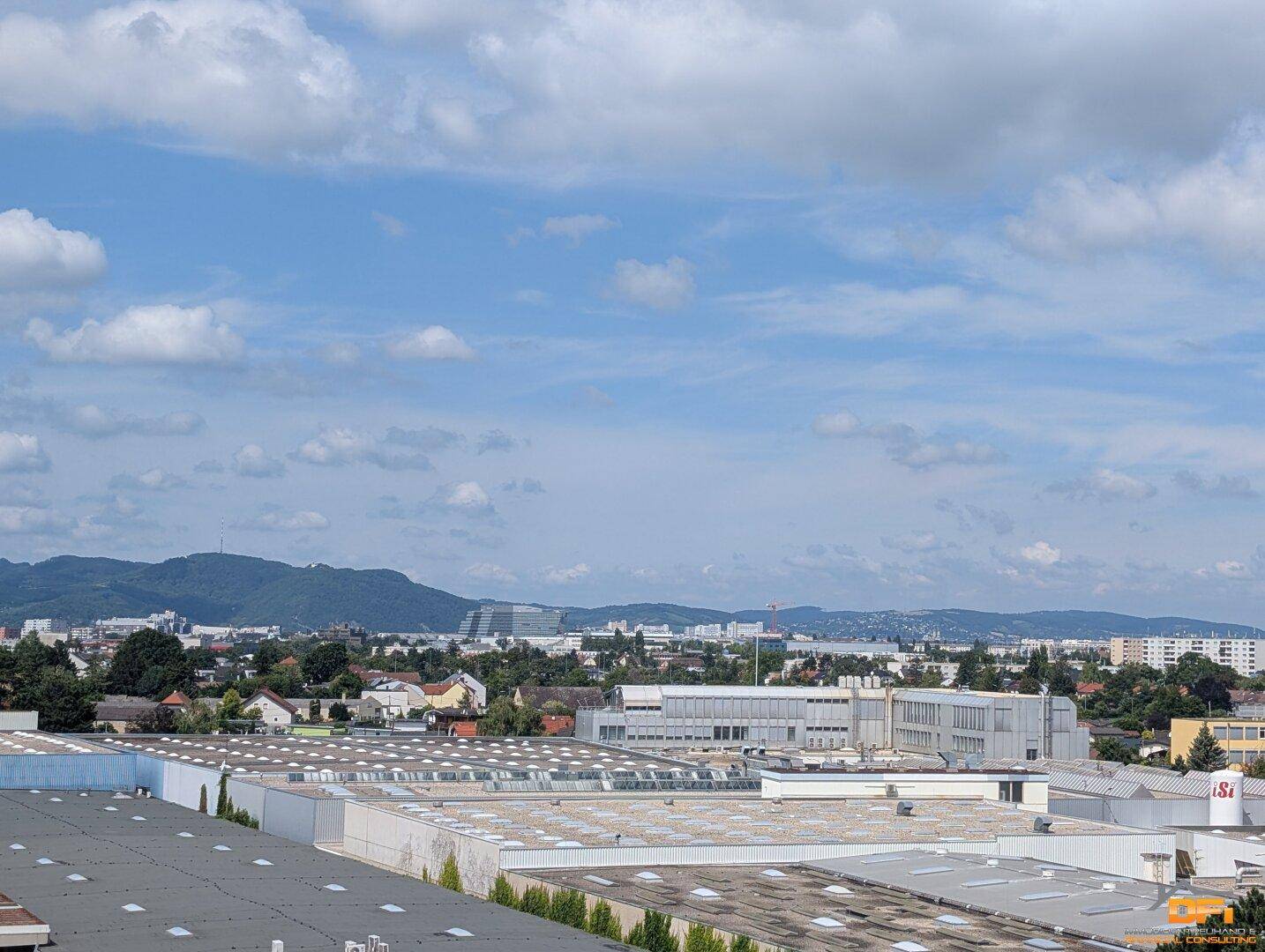 Weitläufiger Blick über eine städtische Landschaft mit Industriegebäuden und Bergen im Hintergrund.