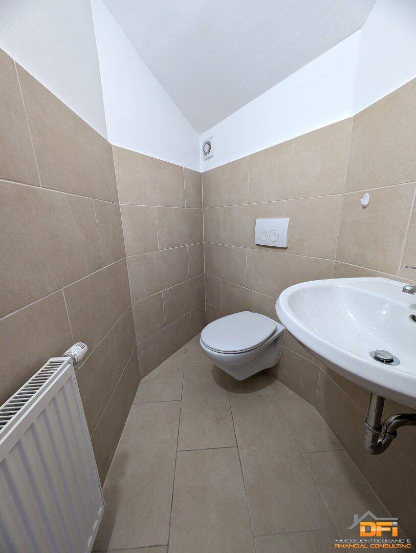 Kompaktes Gäste-WC mit Wand-WC, kleinem Waschbecken und Heizkörper, gefliest in Beige.