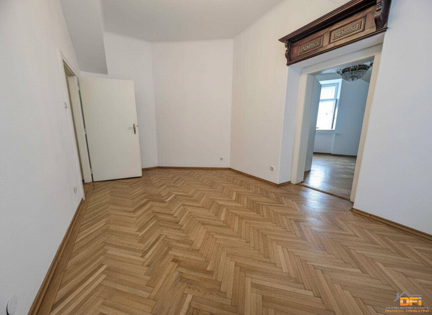 Geräumiges Zimmer mit Fischgrätparkett und einem eleganten Türrahmen, der zu einem anderen Bereich führt.