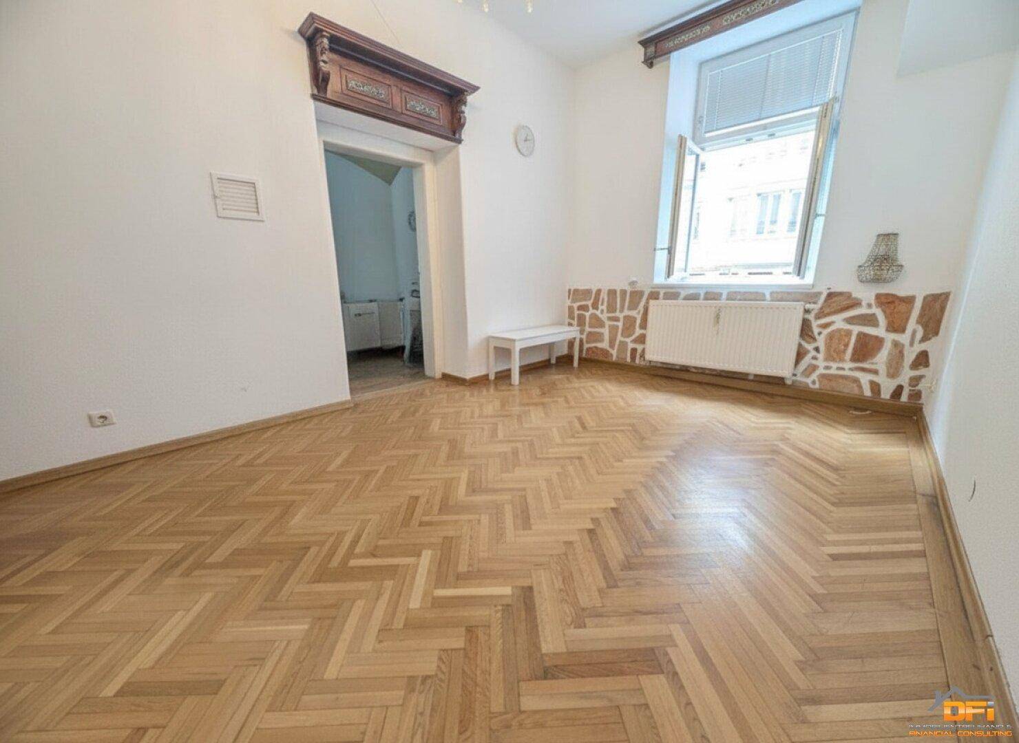 Helles Zimmer mit Fischgrätparkett, dekorativer Wandverkleidung und einem Durchgang zu einem weiteren Raum.