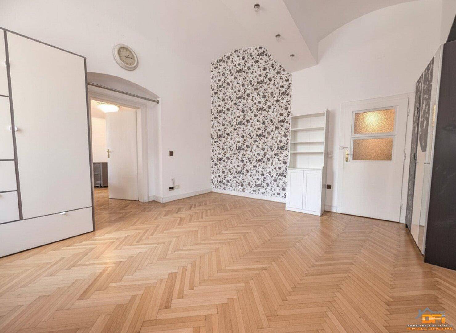 Geräumiges Zimmer mit Fischgrätparkett, einer Akzentwand mit Tapete und einem Durchgang zu einem weiteren Raum.