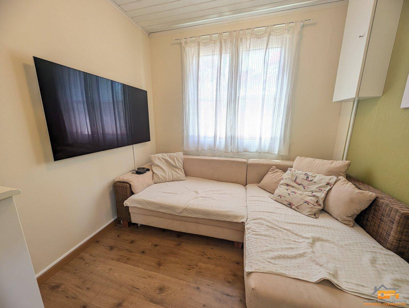 Gemütliches Wohnzimmer mit Sofa, Fernseher und Fenster mit Vorhängen.