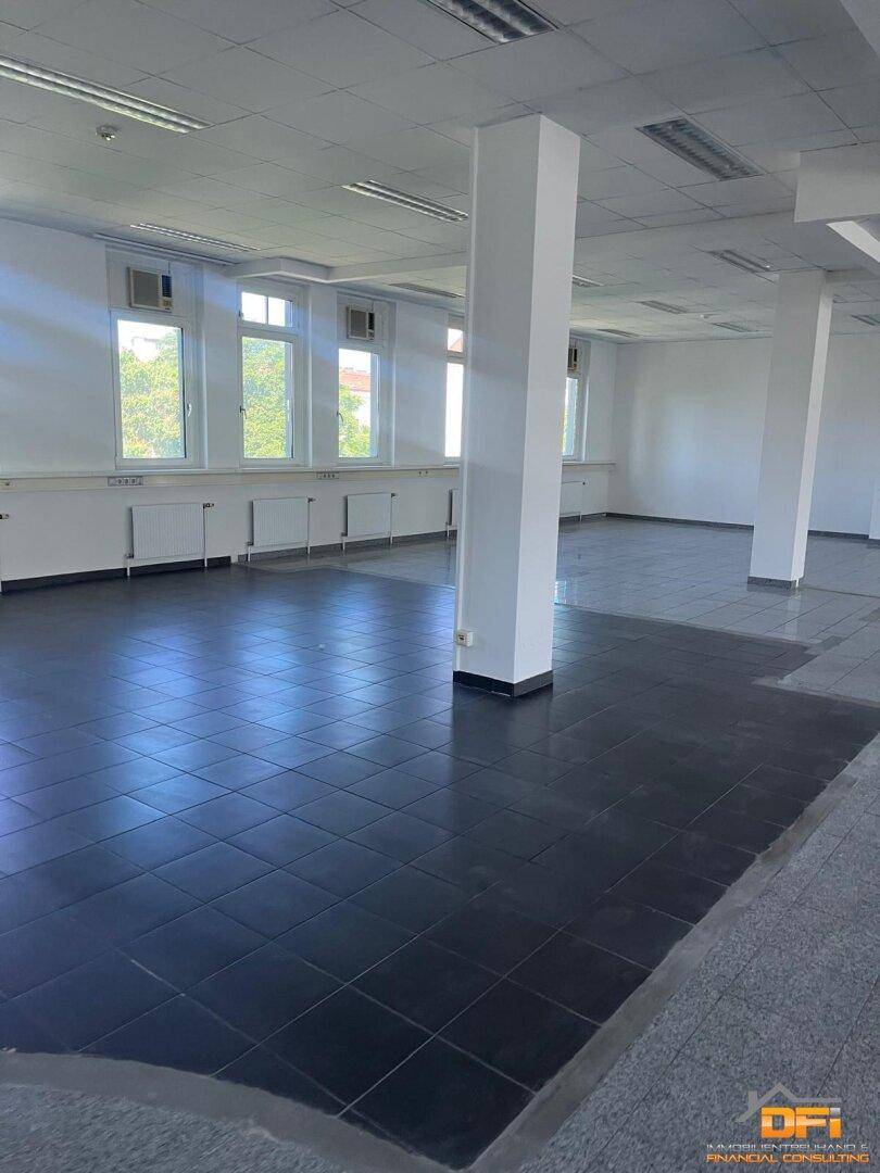 Heller Büroraum mit Säulen, Fliesenboden und Fenstern, die viel Tageslicht hereinlassen.