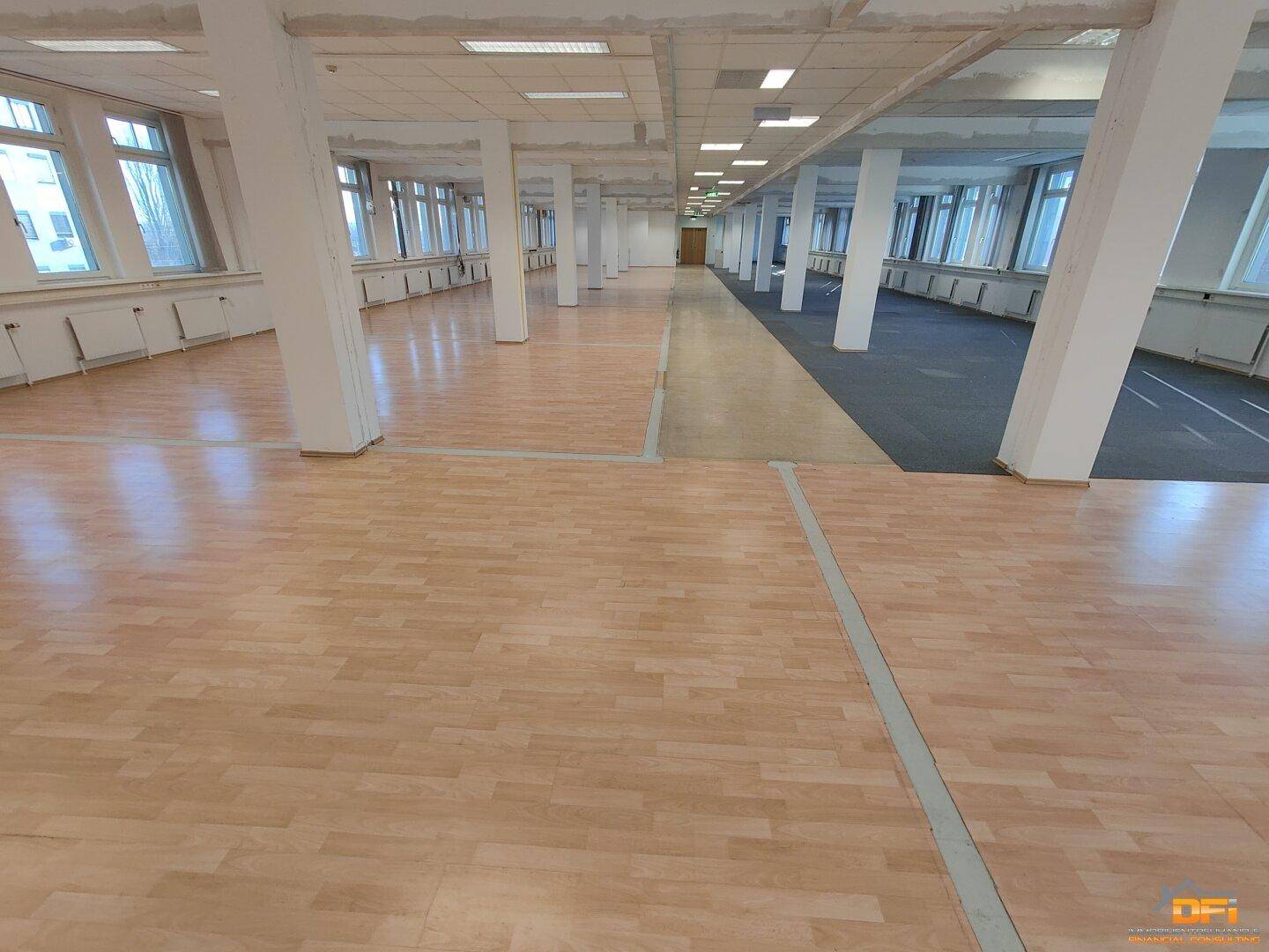 Weitläufiger Bürobereich mit hellem Laminatboden, vielen tragenden Säulen und durchgehenden Fensterfronten.