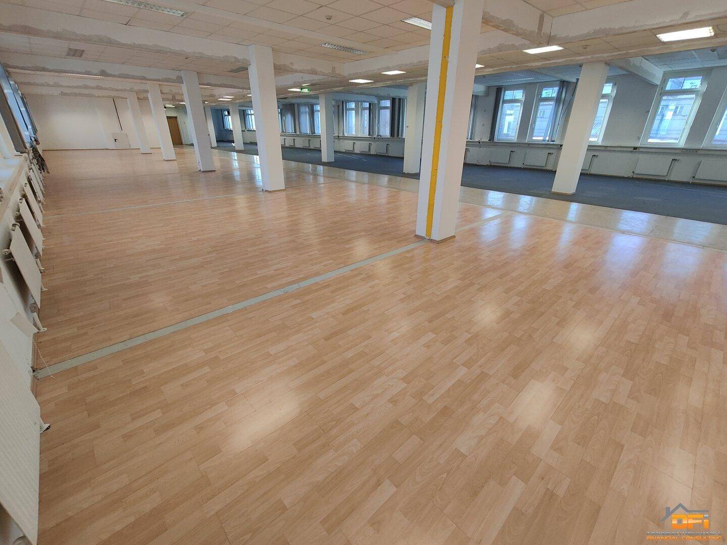 Heller Büroraum mit Holzboden, weißen Säulen und großen Fenstern, die viel Tageslicht hereinlassen.
