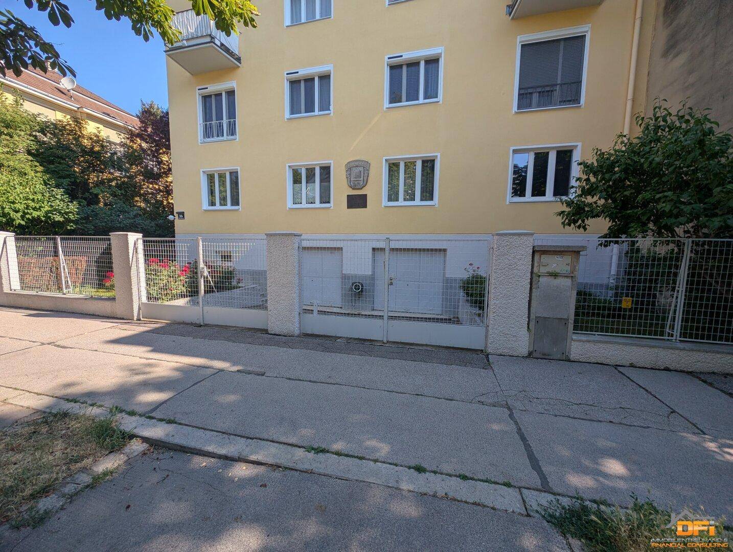 Außenansicht eines Gebäudes mit gelber Fassade, Zaun und Einfahrt zu den Garagen.