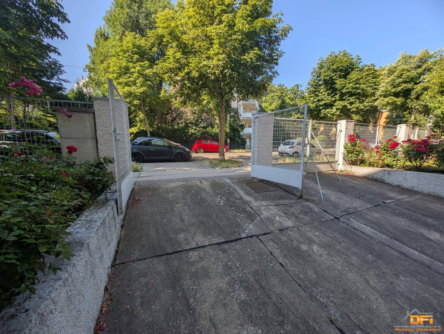 Offenes Einfahrtstor zu einem Grundstück mit Bäumen und parkenden Autos im Hintergrund.