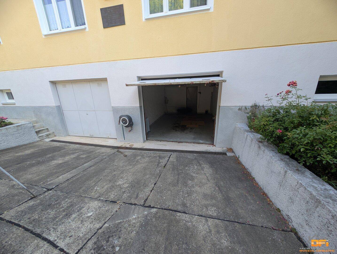 Gebäudefassade mit einer offenen und einer geschlossenen Garagentür sowie gepflasterter Einfahrt.