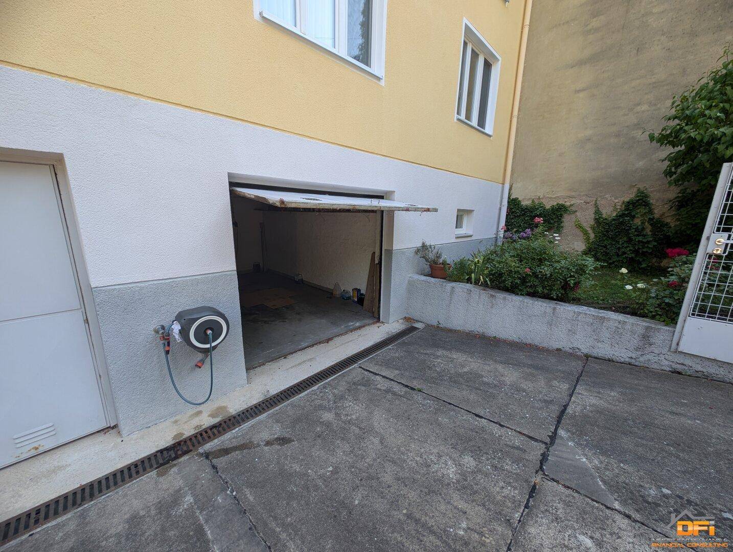 Offene Garagentür mit einem Schlauchaufroller an der Wand und gepflasterter Einfahrt.