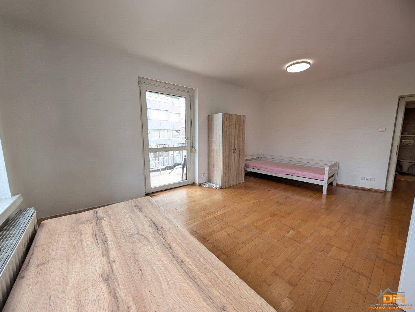 Schlafzimmer mit Parkettboden, Balkonzugang und einfacher Möblierung.