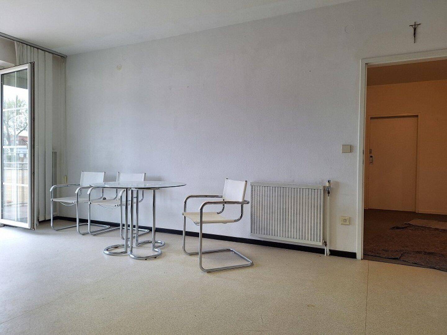 Wohnbereich mit modernem Glastisch und Stühlen, Heizkörper und Zugang zu einem Außenbereich.