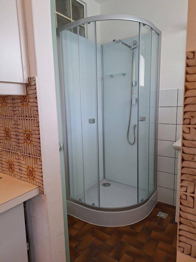 Duschkabine mit Glaswänden in einem Badezimmer, umgeben von weißen Fliesen und Terrakottaboden.