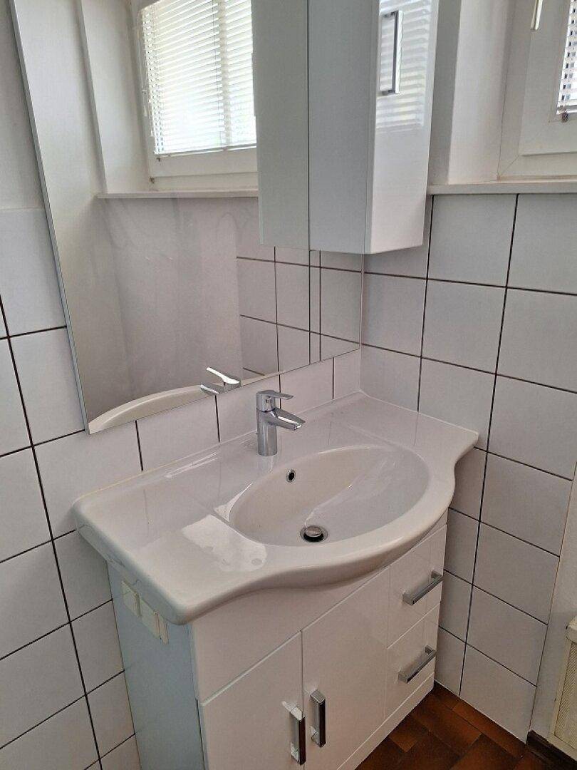 Waschbecken mit Unterschrank und Spiegel in einem hell gefliesten Badezimmer mit Fenster.