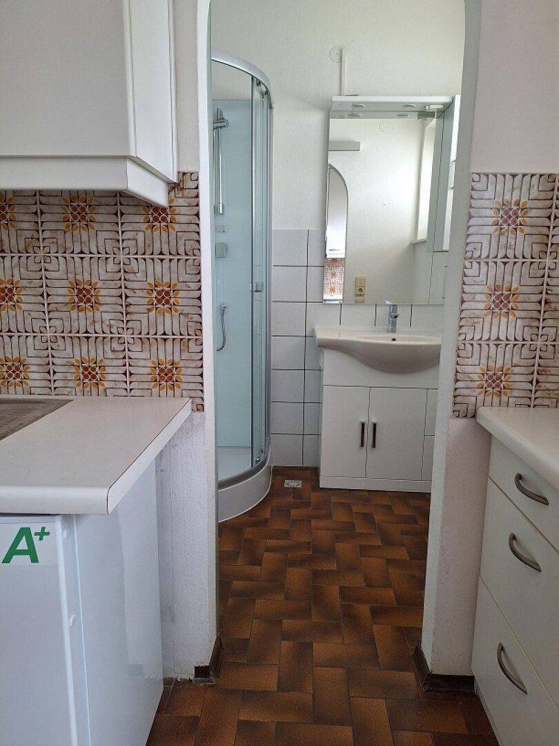 Blick von der Küche in ein Badezimmer mit Duschkabine und Waschbecken, getrennt durch eine Tür.