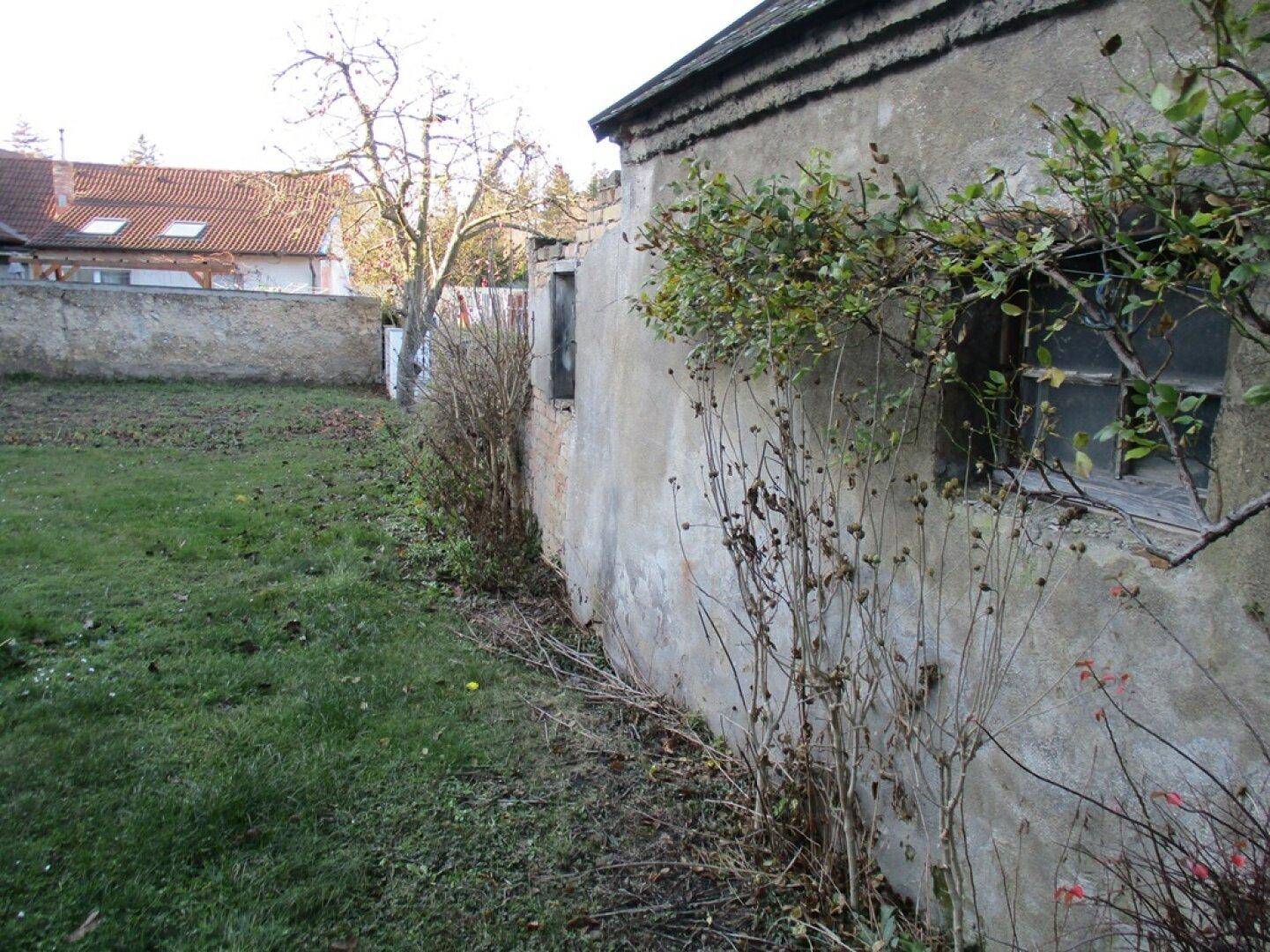 Altes Nebengebäude mit verputzter Fassade und kleinen Fenstern, angrenzend an einen ungepflegten Garten.