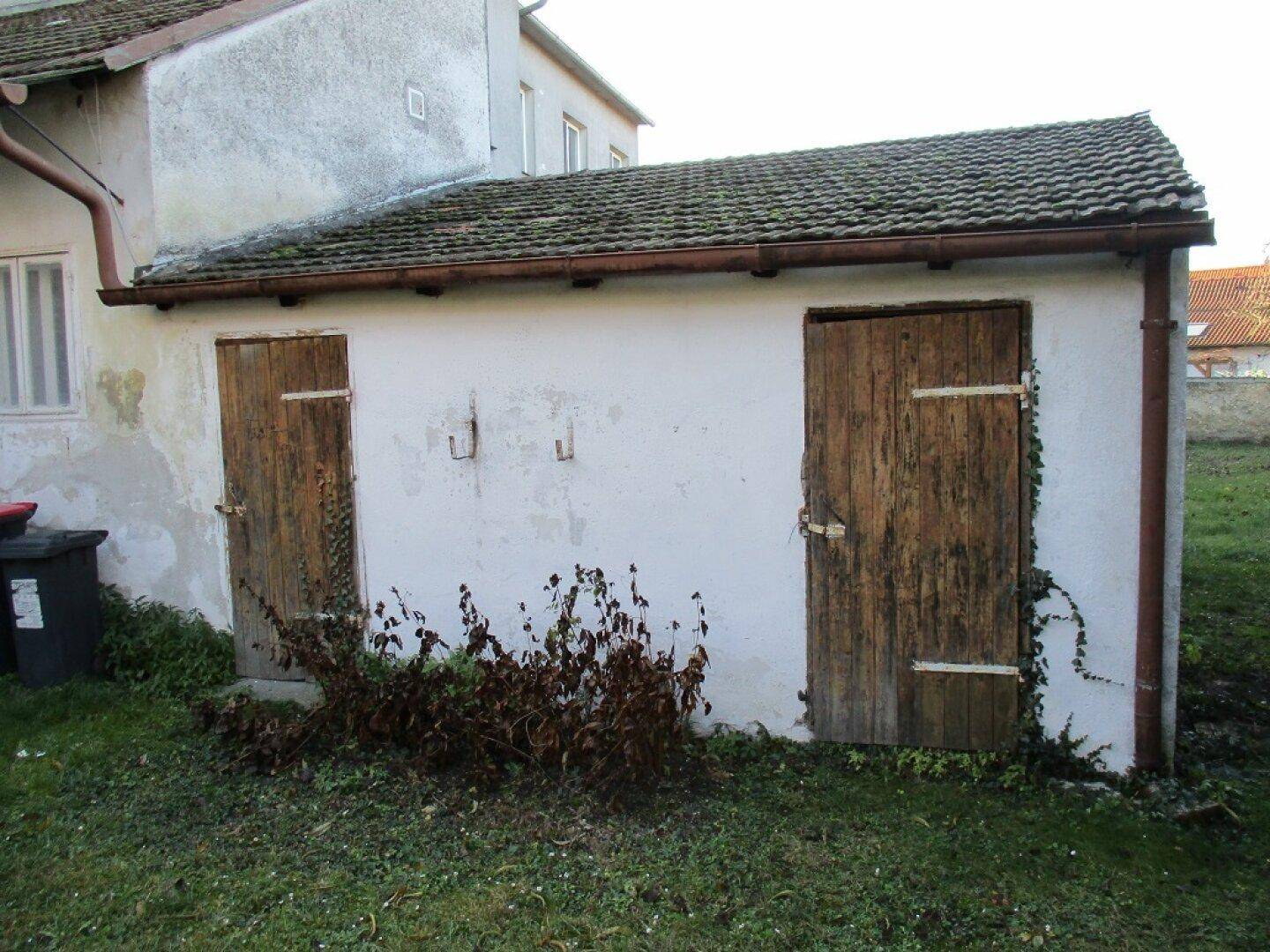 Ansicht des kleinen Nebengebäudes mit rustikalen Holztüren und Ziegeldach im Gartenbereich.