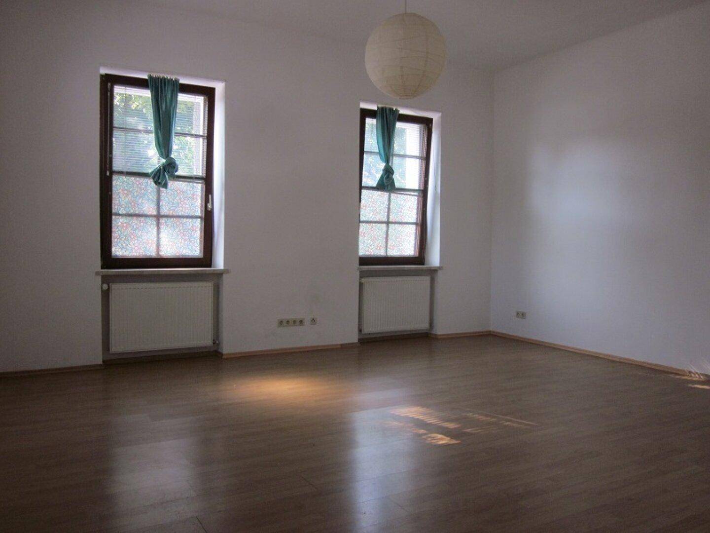 Heller, leerer Raum mit Holzboden, zwei Fenstern und einer Deckenleuchte.
