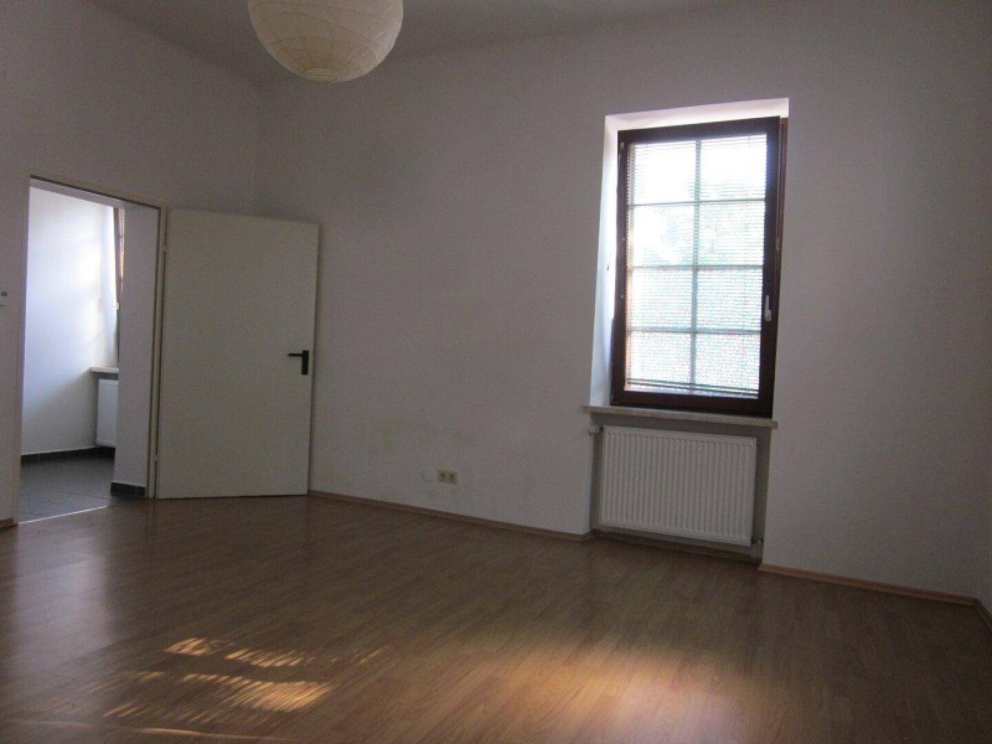 Leerer Raum mit Holzboden, einem Fenster und Zugang zu einem weiteren Zimmer.
