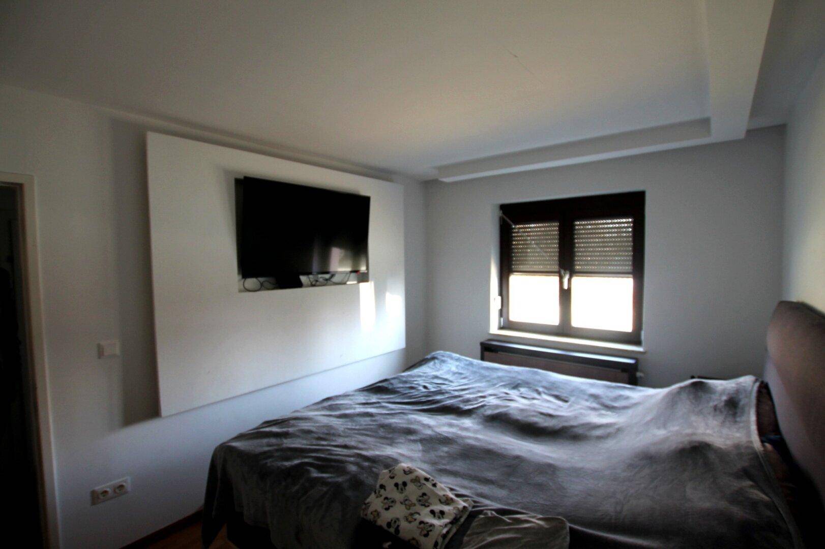 Schlafzimmer mit Doppelbett, Wandfernseher und Fenster mit Rollladen für Privatsphäre.