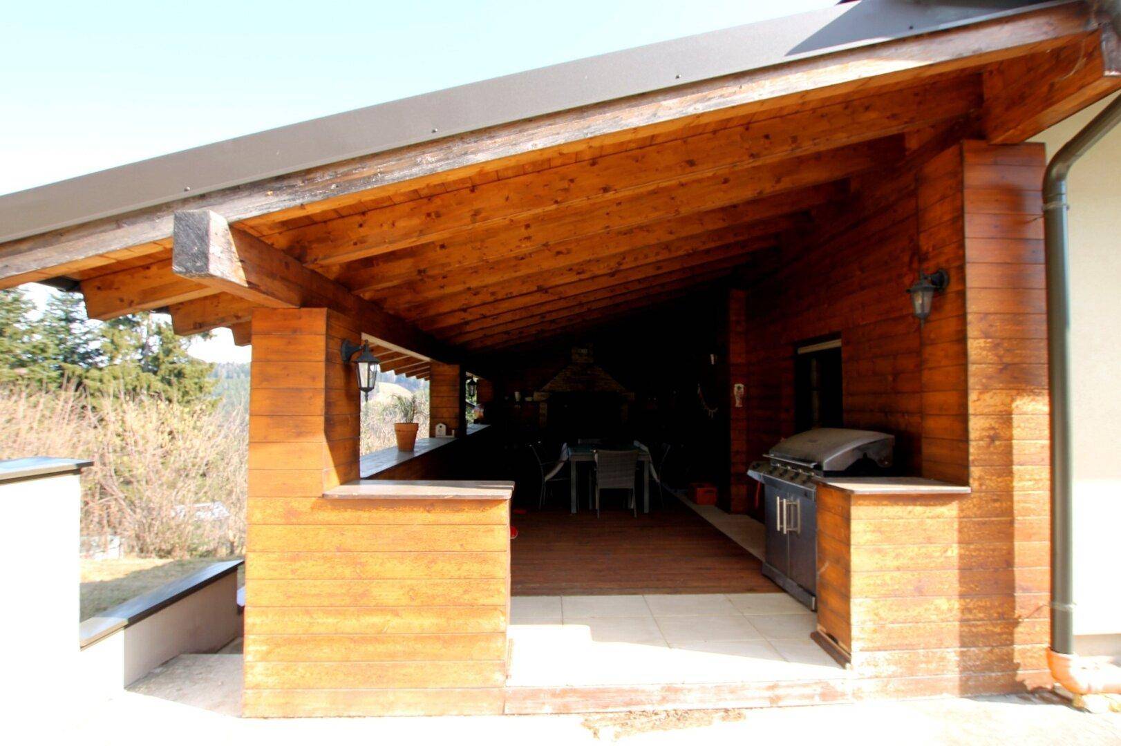 Überdachte Holzterrasse mit Grillbereich und Blick in die umliegende Natur und Bäume.