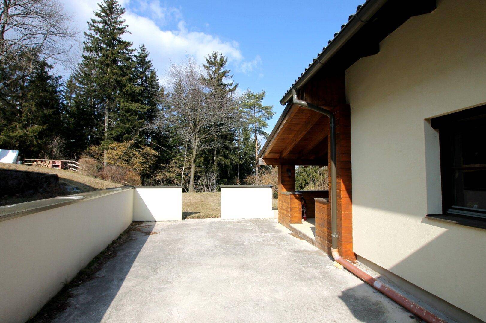 Betonierte Terrasse neben dem Haus mit Zugang zum überdachten Bereich und Garten.