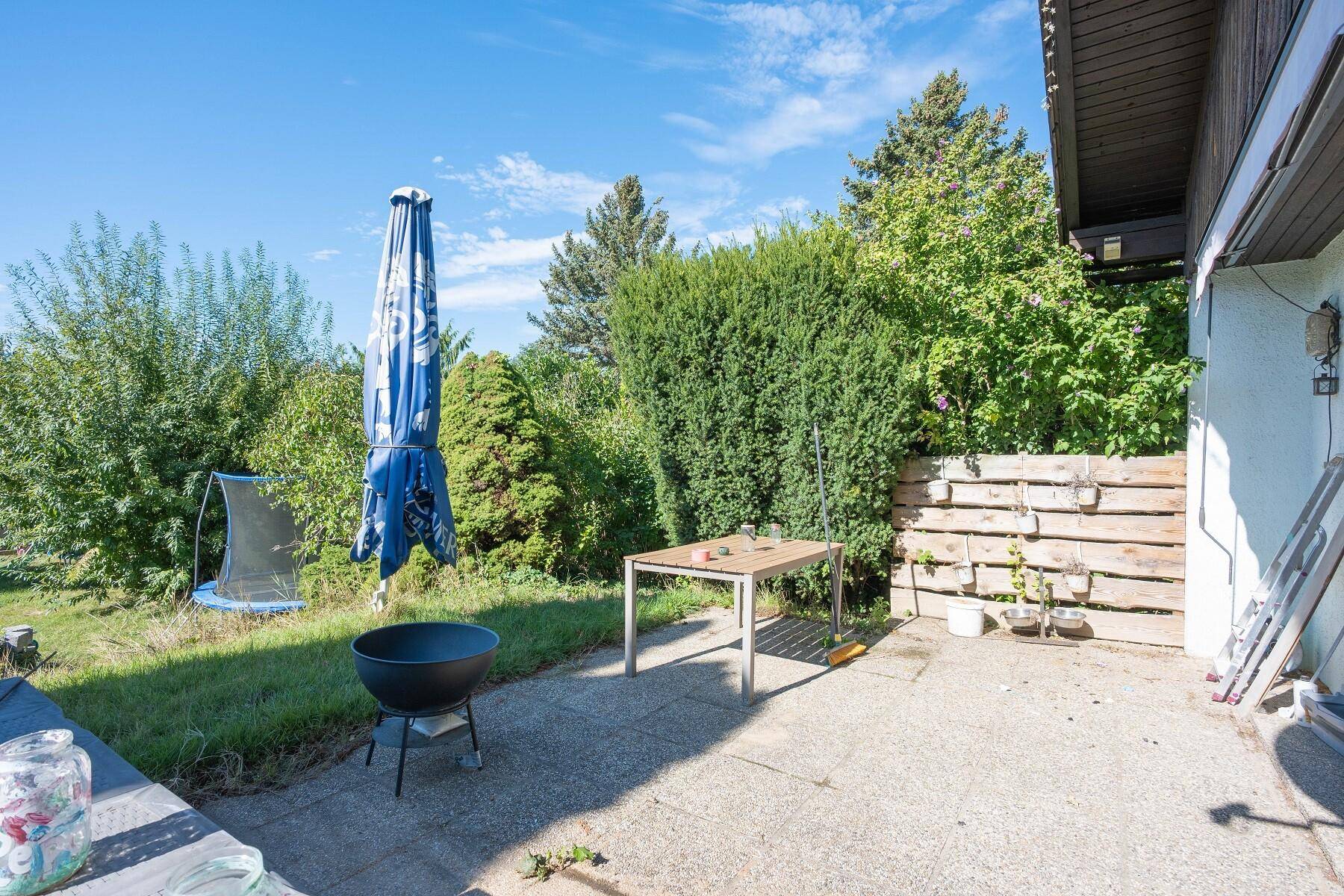 Sonniger Terrassenbereich mit Grill, Gartentisch und Schirm, umgeben von Hecken.