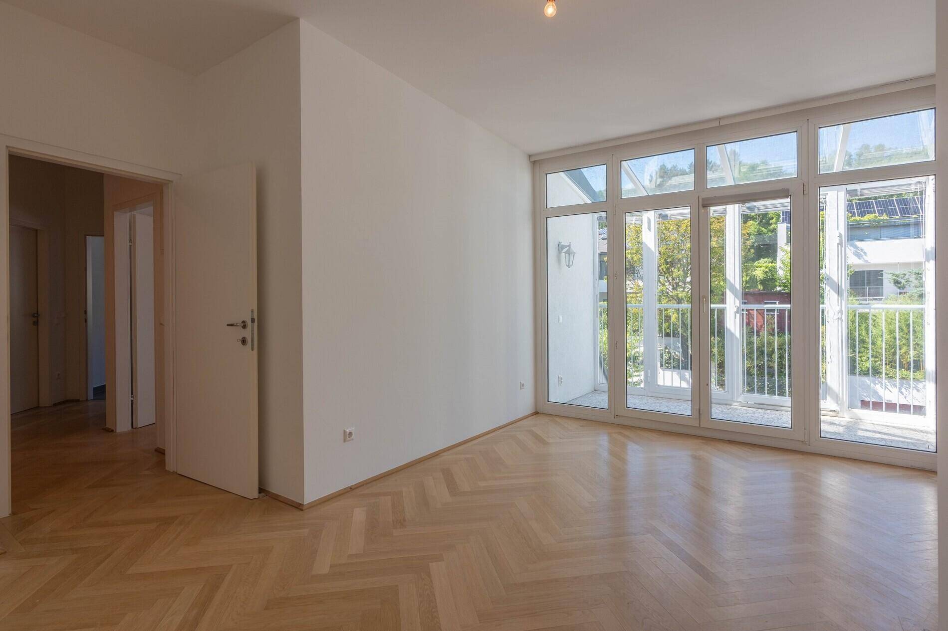 Lichtdurchfluteter Wohnbereich mit Fischgrätparkett und Zugang zu einem verglasten Balkon.