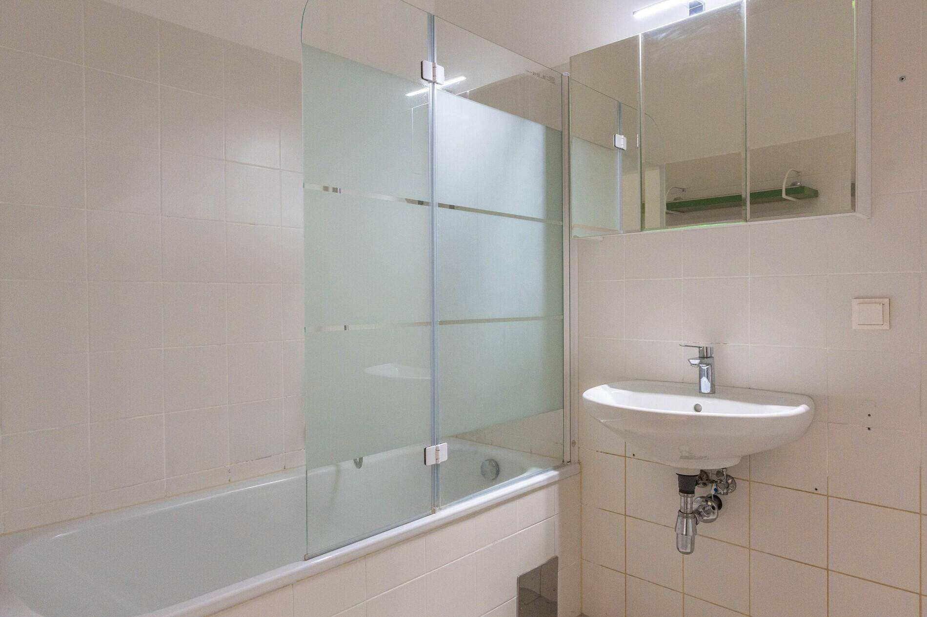 Badezimmer mit weißen Fliesen, Badewanne mit Duschwand und Waschbecken mit Spiegelschrank.
