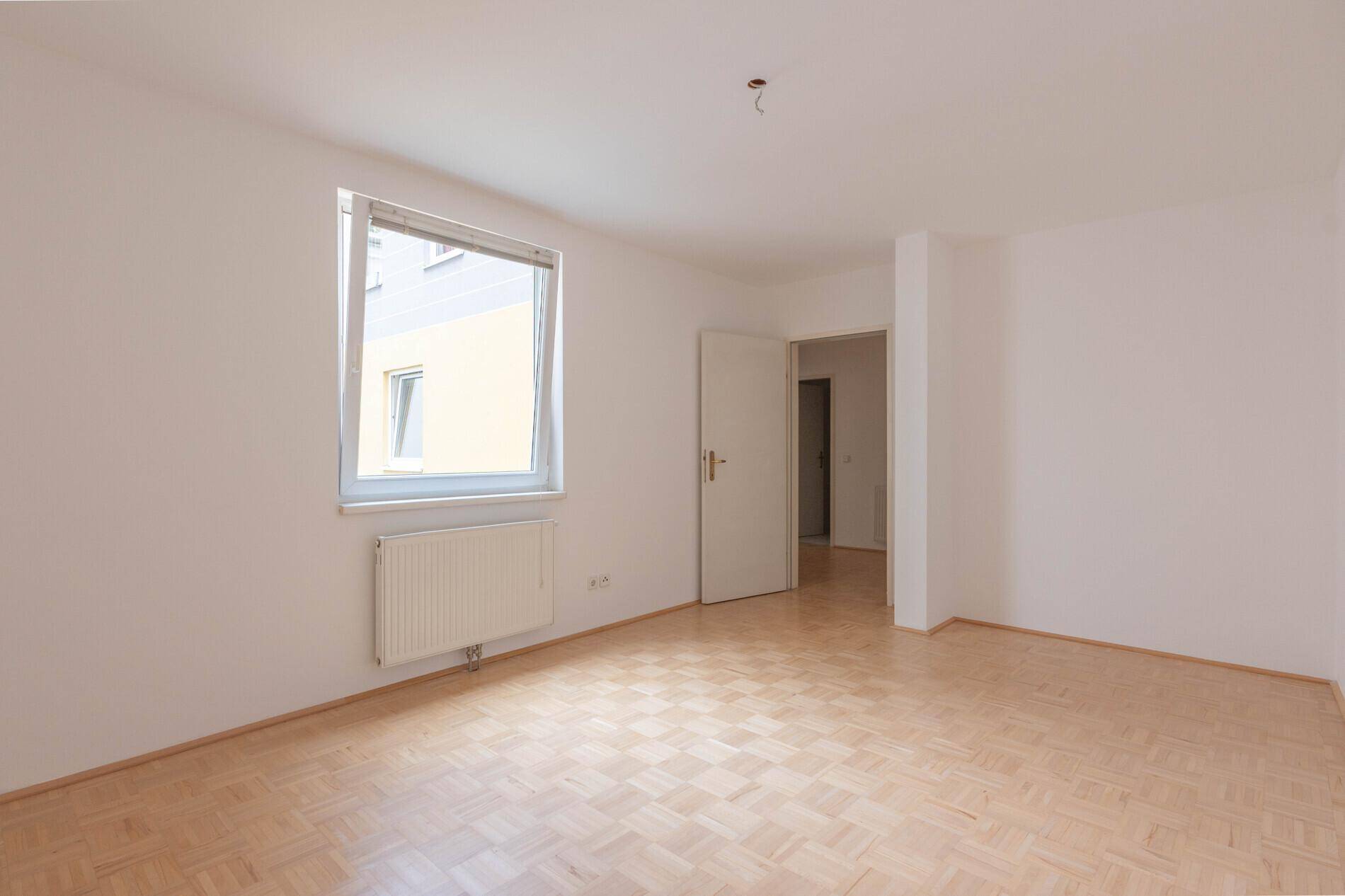Leeres Schlafzimmer mit hellem Parkettboden und einem Fenster, das viel Tageslicht hereinlässt.