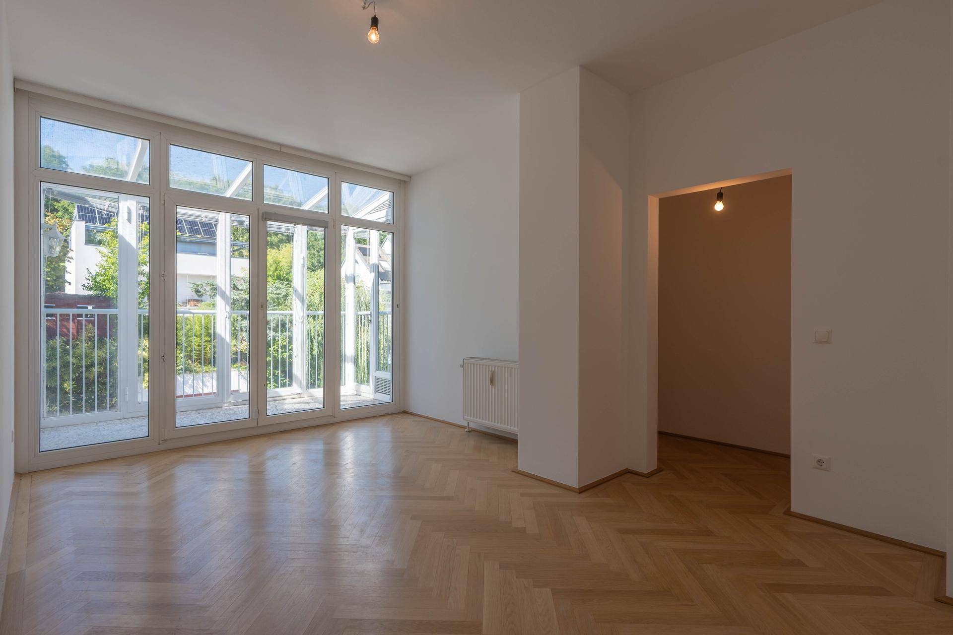Geräumiges Wohnzimmer mit Fischgrätparkett und großen Fenstern, die viel Tageslicht hereinlassen.