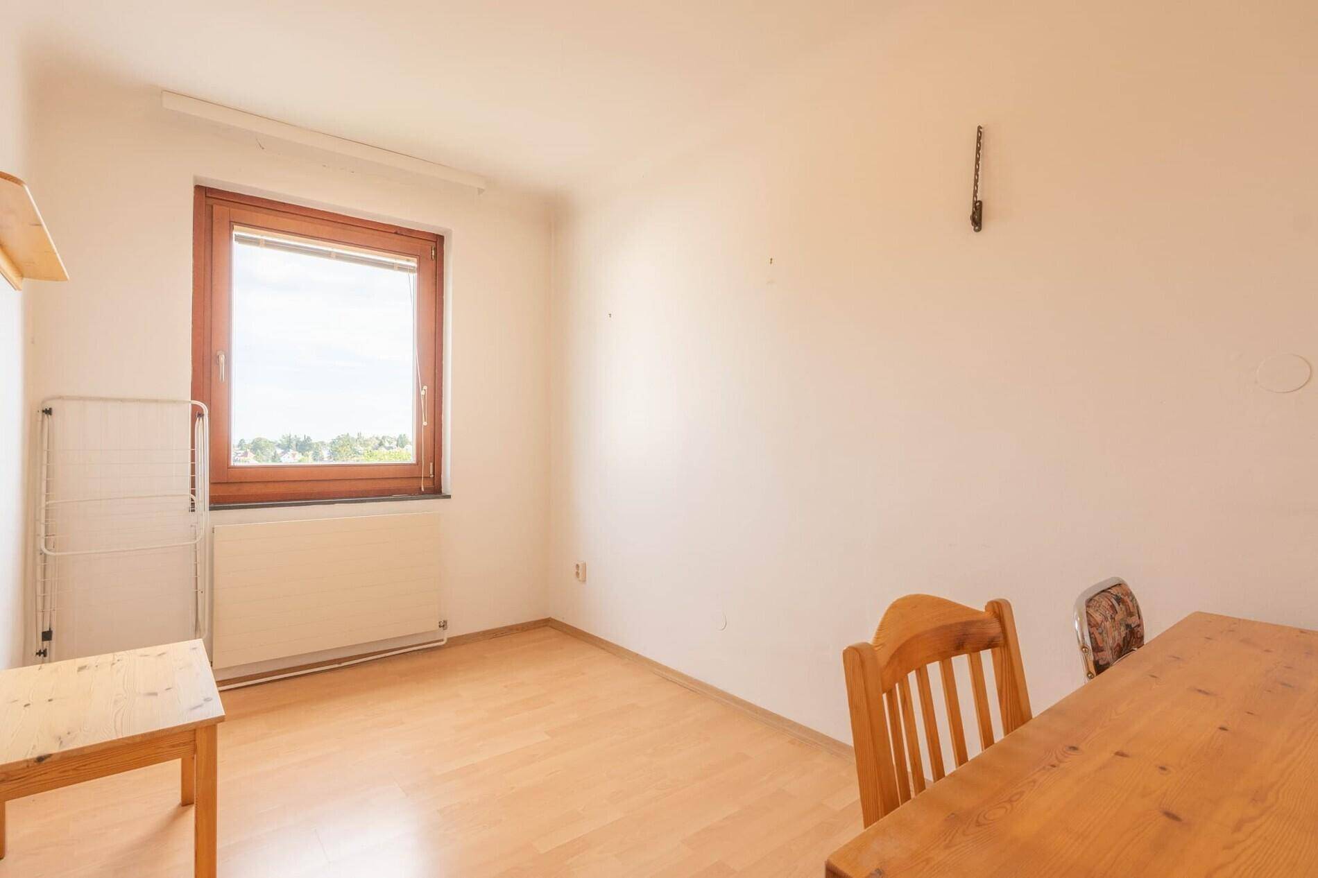 Leeres Zimmer mit Laminatboden, Holztisch und einem Fenster mit Ausblick.