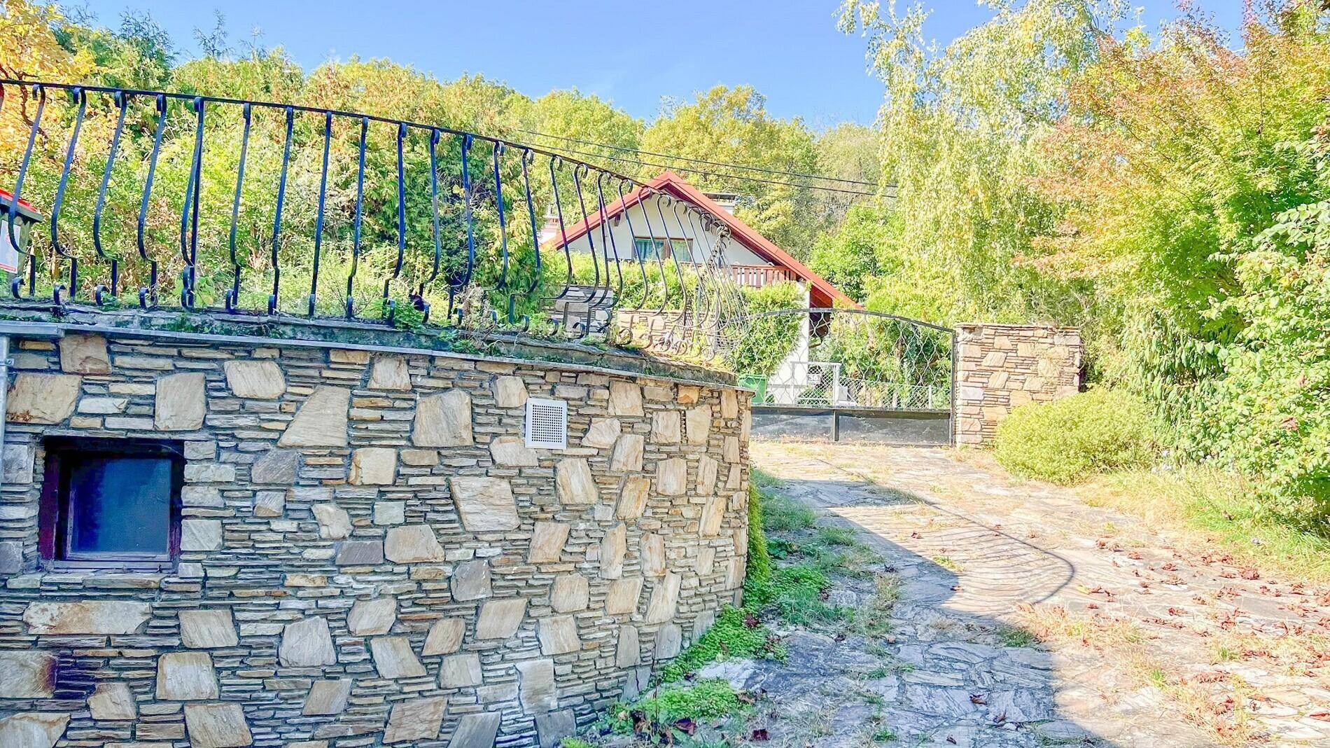 Zufahrt zum Anwesen mit Natursteinmauer, schmiedeeisernem Tor und üppiger Bepflanzung.