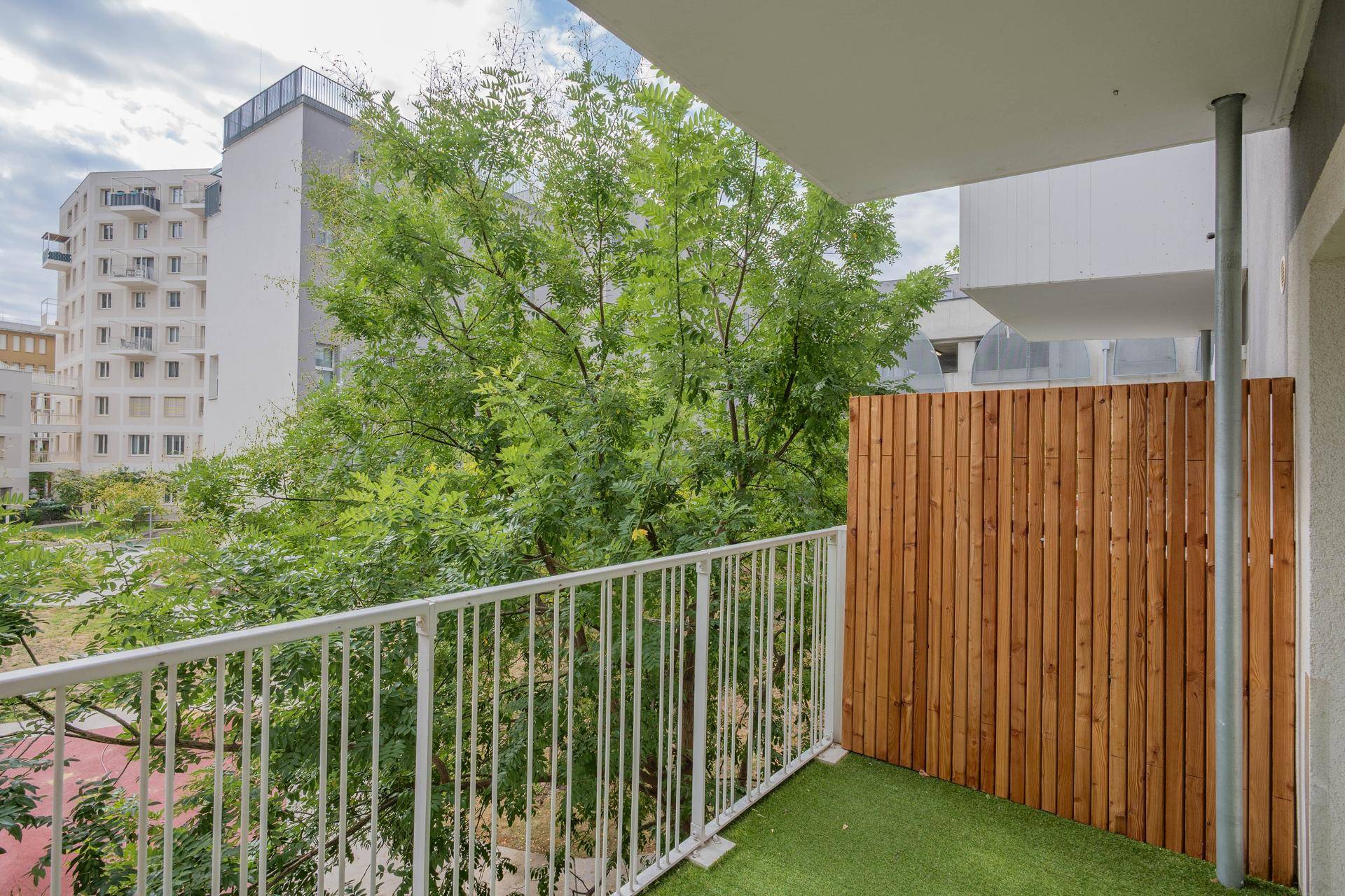 Balkon mit Kunstrasen und Holzsichtschutz, umgeben von Bäumen und modernen Gebäuden.